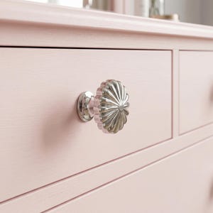Puede incluir: Un pomo de cajón redondo de metal plateado con un patrón estriado similar a un rayo de sol, fijado a un frente de cajón de madera de color rosa claro. El pomo presenta un acabado pulido y reflectante con crestas que irradian hacia el borde.