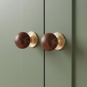 May include: Two spherical cabinet knobs featuring dark brown wood grain heads attached to circular brushed brass bases. The knobs are mounted on a sage green paneled surface, showcasing a smooth finish and a rounded, symmetrical design.