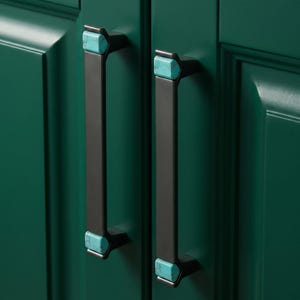 May include: Two vertical cabinet pulls featuring a matte black finish with rectangular turquoise stone inlays at the top and bottom ends. The hardware is mounted on a dark green cabinet door with a raised panel design and a smooth finish.