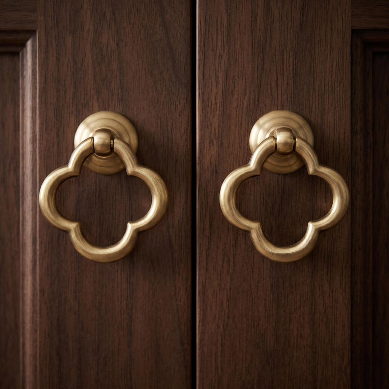 May include: Two brass-toned metal cabinet pulls with a clover-shaped ring design. Each pull features a circular mounting base and a hinged, four-lobed ring handle attached to a dark brown wooden cabinet door with a visible vertical grain pattern.