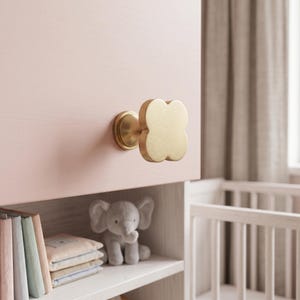 May include: A brushed gold cabinet knob with a four-lobed clover shape attached to a light pink wooden surface. The knob features a smooth, matte metallic finish and a circular base plate, providing a functional handle for furniture doors.