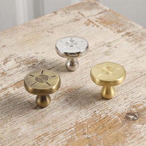May include: Three metal cabinet knobs with a mushroom-shaped head and a central spiral sun design. One knob has a polished silver finish, one has a brushed antique brass finish, and one has a smooth matte gold finish.