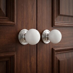 May include: Two round white porcelain door knobs with polished silver-toned metal bases. Each knob is mounted on a dark brown wooden door with a paneled design. The hardware features a smooth, spherical handle attached to a circular backplate.