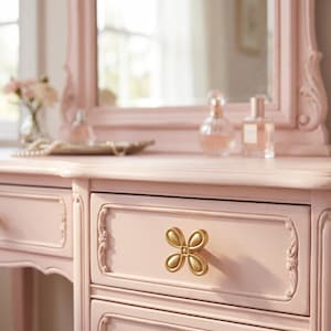 May include: A light pink vanity table featuring a central drawer with a gold-toned, four-petaled flower-shaped knob. The furniture piece has carved decorative molding along the drawer edges and a matching pink mirror frame positioned above the surface.