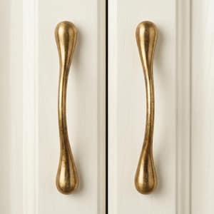 May include: Two vertical cabinet pulls with a curved, elongated teardrop shape and a brushed antique gold finish. The handles are mounted on a white paneled door surface, featuring a smooth, metallic texture with subtle variations in tone.