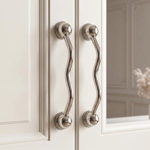 May include: Two polished silver-toned metal cabinet handles with a distinct wavy, serpentine silhouette. Each handle features rounded, spherical mounting bases at the top and bottom, securely attached to a light-colored, paneled cabinet door surface.