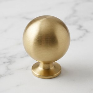 Puede incluir: Un pomo esf&eacute;rico para muebles con acabado en oro cepillado. El pomo presenta una superficie superior lisa y redondeada que se transforma en una base cil&iacute;ndrica c&oacute;nica, conectada a una placa de montaje circular plana en la parte inferior.