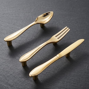 May include: Three gold-colored metal cabinet pulls shaped like miniature cutlery. The set includes one spoon-shaped pull, one fork-shaped pull, and one knife-shaped pull, each featuring a polished finish and two small cylindrical mounting posts.
