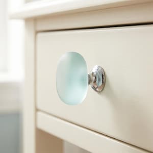 May include: A round, frosted light blue glass cabinet knob with a polished silver-tone metal base. The knob is attached to a cream-coloured wooden drawer front, featuring a smooth, matte finish and a simple, circular, translucent design.