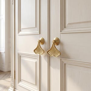 May include: Two gold-toned metal door handles with a fan-shaped design and vertical ribbed detailing, mounted on a pair of off-white double doors featuring recessed rectangular panels and a visible wood grain texture on the door surface.