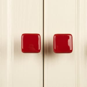 May include: Two square cabinet knobs with a glossy, solid red finish. Each knob features rounded corners and a smooth, reflective surface, mounted side-by-side on a vertical cream-colored paneled surface with a central seam running between them.