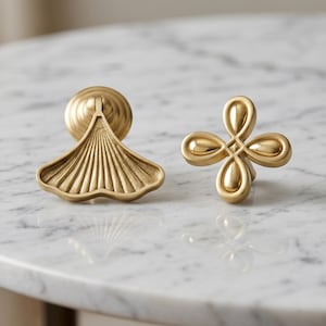 May include: Two gold-toned metal cabinet knobs. One knob features a fan-shaped design with vertical ridges, while the other knob has a symmetrical four-petaled floral shape with smooth, rounded edges and a central square detail.