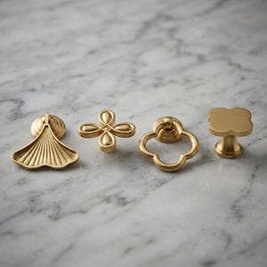 May include: Four distinct brass-toned metal cabinet knobs and pulls arranged in a row. The set includes a fan-shaped drop pull, a four-petaled floral knob, a rounded ring pull with a clover-like frame, and a solid four-lobed clover knob.