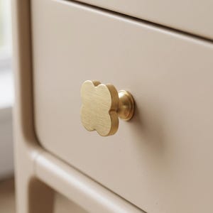 May include: A brushed brass cabinet knob shaped like a four-leaf clover attached to a light beige drawer front. The metal hardware features a smooth, matte finish with rounded edges, providing a functional grip for opening the furniture piece.