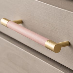 May include: A drawer pull handle featuring a cylindrical center wrapped in a soft pink textured material, flanked by two polished gold-toned metal end caps and mounting posts, attached horizontally to a light gray wooden drawer front surface.