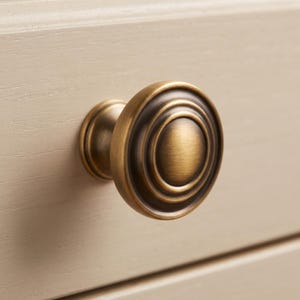 以下が含まれることがあります： ブラッシュドアンティーク真鍮仕上げの丸いキャビネットノブ。ノブは、中央の盛り上がったドームと段差のあるエッジを備えた同心円状のデザインが特徴で、層状の外観を作り出しています。金属表面の溝には暗いハイライトがあります。