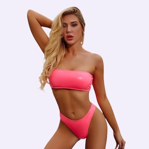 Puede incluir: Un conjunto de bikini de dos piezas en color rosa neón brillante que presenta un top tipo bandeau sin tirantes y una braga de bikini de corte alto. El traje de baño está hecho de un material liso de color sólido con un acabado mate.