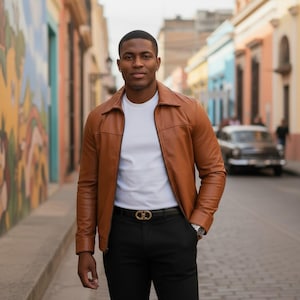 May include: A tan leather jacket worn over a plain white t-shirt, paired with black trousers and a black belt featuring a gold-toned buckle. The jacket has a classic collar, long sleeves, and a front zipper closure, creating a clean, layered look.