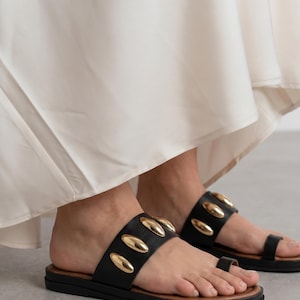 May include: Black leather sandals with gold-colored oval studs. The sandals have a brown sole and a toe loop. The image shows the sandals being worn with a white dress.