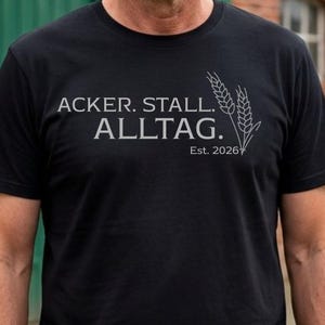 Könnte beinhalten: Schwarzes Rundhals-T-Shirt mit dem Text "ACKER. STALL. ALLTAG." in Hellgrau. Eine Grafik von Weizenähren befindet sich rechts vom Text. Der Text "Est. 2026" steht unter dem Wort "ALLTAG."