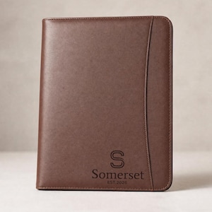 May include: A dark brown rectangular portfolio with a smooth finish and stitched edges. The front features a vertical curved seam on the right side and the embossed text "S Somerset EST.2026" centred in the lower portion of the cover.