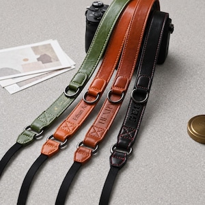 Può includere: Quattro tracolle per fotocamera in pelle verde oliva, marrone chiaro, cuoio e nero. Ogni tracolla presenta anelli in metallo, fettuccia in nylon nero e testo in rilievo personalizzato, inclusi Ethan Adams, HENRY e GEO. Le tracolle sono fissate a una fotocamera.