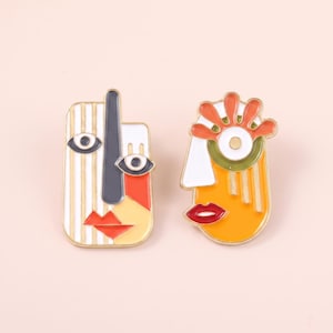 May include: Two enamel pins featuring abstract face designs with gold-toned metal outlines. The left pin is rectangular with white stripes, a grey nose, and red lips. The right pin is rounded with orange, white, and green details and red lips.