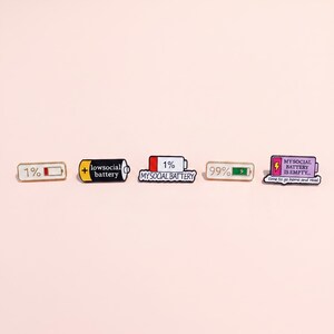 Könnte beinhalten: Fünf Emaille-Anstecknadeln mit Batterie-Motiven. Die Anstecknadeln zeigen verschiedene Batteriestände und Texte, darunter 1%, low social battery, 99% und ein lila Batteriesymbol mit dem Text MY SOCIAL BATTERY IS EMPTY... time to go home and read.