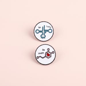 May include: Two circular enamel pins with white backgrounds and black rims. The top pin features a blue molecular structure and the text Be Lucky. The bottom pin displays a chemical structure with a red heart icon and the text Be Happy.