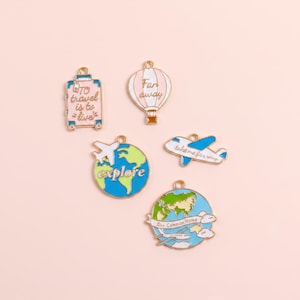 May include: Five metal enamel charms featuring travel themes: a pink suitcase with the text To travel is to live, a pink hot air balloon with Far away, a globe with a plane and explore, a blue airplane, and a globe with Our Common Home.