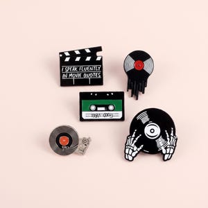 Könnte beinhalten: Ein Set aus fünf Emaille-Anstecknadeln bestehend aus einer schwarzen Filmklappe mit dem Text I SPEAK FLUENTLY IN MOVIE QUOTES, einer schmelzenden schwarzen Schallplatte, einer grün-schwarzen Kassette, einer Schallplatte auf einem Plattenspieler und einer Schallplatte mit Skeletthänden.