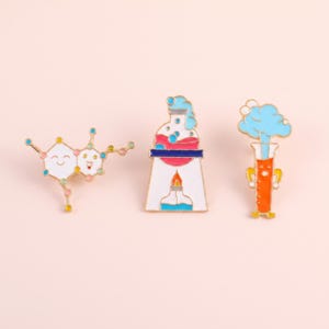 May include: Three enamel pins featuring science themes: a white molecular structure with smiling faces, a white laboratory flask over a flame with pink liquid and blue bubbles, and an orange test tube with a blue cloud and a smiling face.
