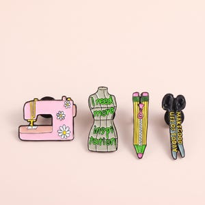 May include: Four enamel pins featuring sewing themes: a pink sewing machine with white daisies, a dress form with the text I need more inspiration, a yellow pencil with a zipper, and black scissors with the text MAKE GOOD USE OF TODAY.