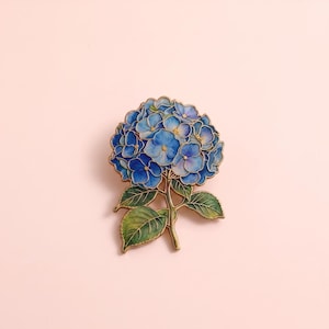 Op de afbeelding: Een metalen broche in de vorm van een hortensiabloem met blauwe en paarse bloemblaadjes, groene bladeren en goudkleurige omlijningen. Het bloemmotief heeft meerdere gelaagde bloemblaadjes en een steel, uitgevoerd in een cloisonné-stijl met emaille.