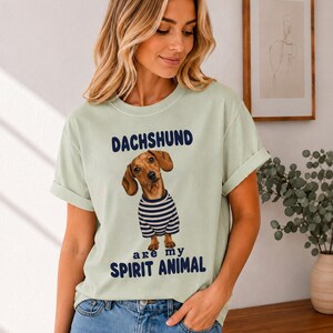May include: A light sage green short-sleeve t-shirt featuring a graphic of a brown dachshund wearing a blue and white striped shirt. The text above the dog reads DACHSHUND and the text below reads are my SPIRIT ANIMAL in dark blue lettering.