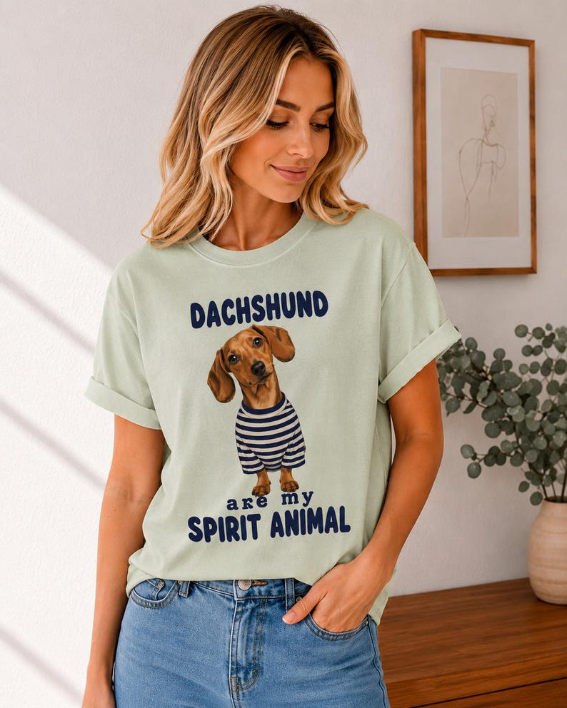 May include: A light sage green short-sleeve t-shirt featuring a graphic of a brown dachshund wearing a blue and white striped shirt. The text above the dog reads DACHSHUND and the text below reads are my SPIRIT ANIMAL in dark blue lettering.