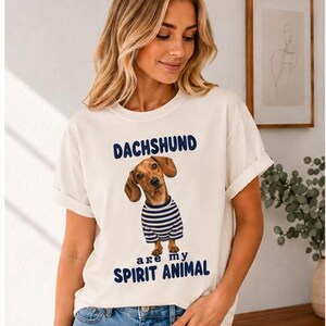 May include: An off-white short-sleeve t-shirt featuring a graphic of a brown dachshund wearing a blue and white striped shirt. The text above the dog reads DACHSHUND and the text below the dog reads are my SPIRIT ANIMAL in blue lettering.