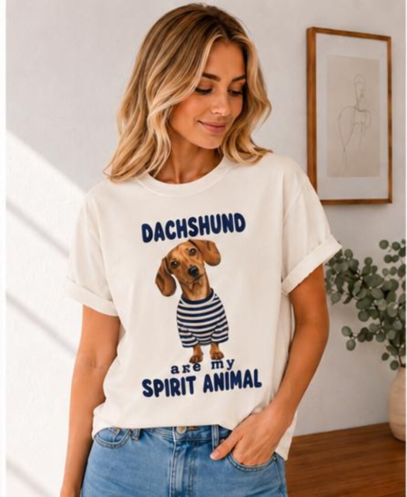 May include: An off-white short-sleeve t-shirt featuring a graphic of a brown dachshund wearing a blue and white striped shirt. The text above the dog reads DACHSHUND and the text below the dog reads are my SPIRIT ANIMAL in blue lettering.