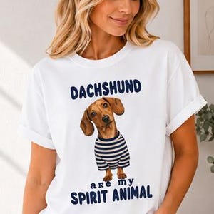May include: A white short-sleeve t-shirt featuring a graphic of a brown dachshund wearing a blue and white striped shirt. The text above the dog reads DACHSHUND and the text below the dog reads are my SPIRIT ANIMAL in dark blue lettering.
