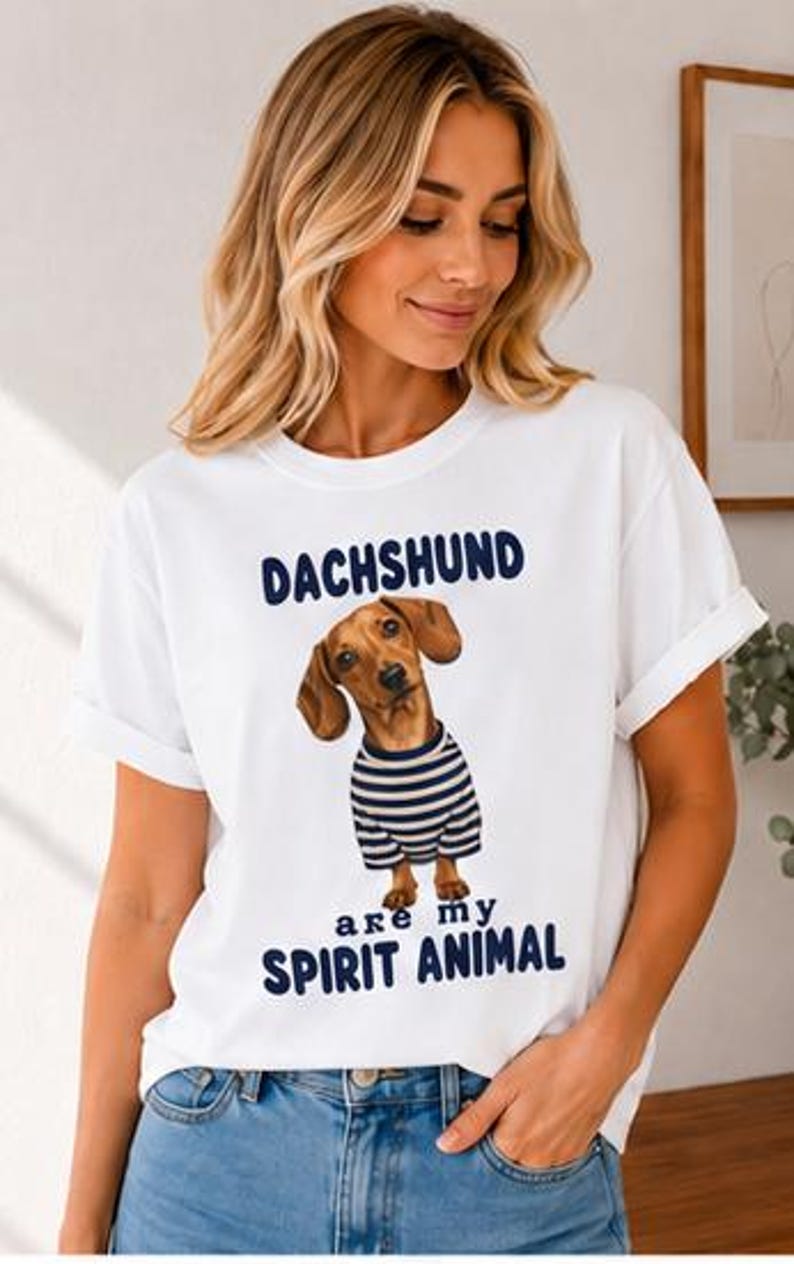 May include: A white short-sleeve t-shirt featuring a graphic of a brown dachshund wearing a blue and white striped shirt. The text above the dog reads DACHSHUND and the text below the dog reads are my SPIRIT ANIMAL in dark blue lettering.