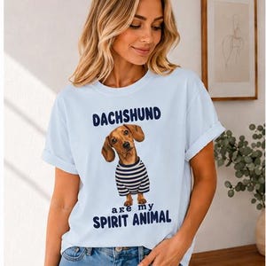 May include: A light blue short-sleeved t-shirt featuring a graphic of a brown dachshund wearing a navy and white striped shirt. The text above the dog reads DACHSHUND and the text below the dog reads are my SPIRIT ANIMAL in dark blue lettering.