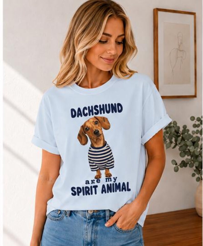 May include: A light blue short-sleeved t-shirt featuring a graphic of a brown dachshund wearing a navy and white striped shirt. The text above the dog reads DACHSHUND and the text below the dog reads are my SPIRIT ANIMAL in dark blue lettering.