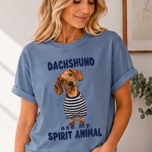 May include: A dusty blue short-sleeve t-shirt featuring a graphic of a brown dachshund wearing a navy and white striped shirt. The text above the dog reads DACHSHUND and the text below reads are my SPIRIT ANIMAL in dark blue lettering.