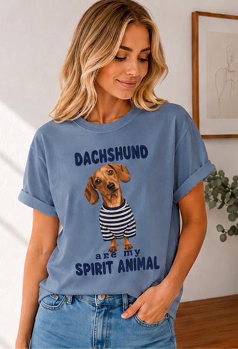 May include: A dusty blue short-sleeve t-shirt featuring a graphic of a brown dachshund wearing a navy and white striped shirt. The text above the dog reads DACHSHUND and the text below reads are my SPIRIT ANIMAL in dark blue lettering.