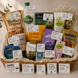 Könnte beinhalten: Ein Geschenkkorb mit verschiedenen Snacks und Pflegeprodukten, darunter Werther's Original Popcorn, Volvic Wasser, Meßmer Kräutertee, Alverde Handcreme und Lippenpflege, Leibniz Butterkekse, Milka Schokolade, Haribo Goldbären, Wasa Knäckebrot, Dallmayr Kaffee und Tic Tac Minzpastillen, alle versehen mit kleinen weißen Kärtchen mit deutschem Text.