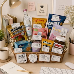 Pode incluir: Uma cesta de presente cheia de vários lanches e bebidas, incluindo Haribo Goldbären, água Gerolsteiner, Leibniz Minis, chocolate Ritter Sport, doces Katjes, chá Meßmer, um produto de bem-estar Balea, gotas Ricola e Nutella, com pequenas etiquetas brancas.