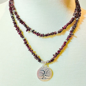 May include: A layered necklace featuring small, deep red garnet stones. The necklace has a silver pendant with a tree of life design. The pendant hangs from the lower layer of the necklace. The necklace is displayed on a white mannequin.