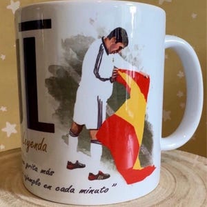 Könnte beinhalten: Ein weißer Keramikbecher mit einer Grafik eines Fußballspielers in weißer Uniform, der eine spanische Flagge hält. Der Text auf dem Becher enthält das Wort Leyenda und den Satz, "un grita más un ejemplo en cada minuto".