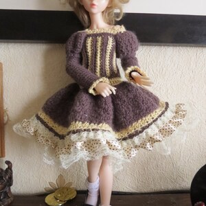 May include: A doll wearing a brown crocheted dress with a yellow and brown striped bodice and a white lace trim. The dress has a brown satin ribbon with white polka dots.