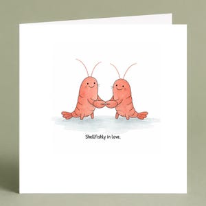 May include: A square greeting card featuring a watercolor illustration of two smiling pink shrimp holding claws. Below the shrimp, the text reads, Shellfishly in love. The card is white with a clean design, showcasing the two creatures standing together.